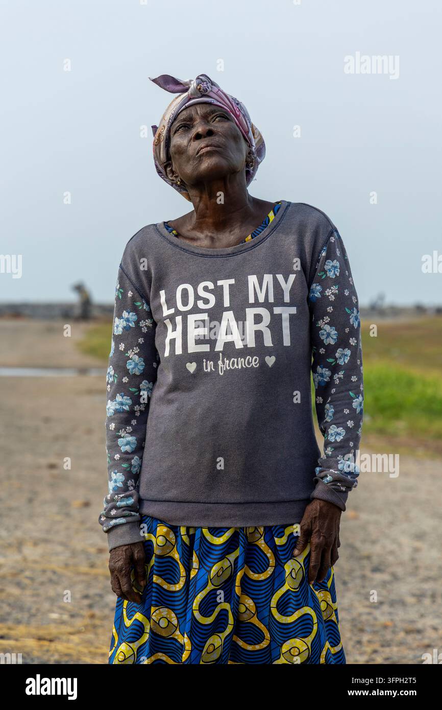 Una donna anziana con un foulard guarda verso l'alto mentre è in piedi su una strada costiera, Regione Centrale, Elmina, Ghana Foto Stock
