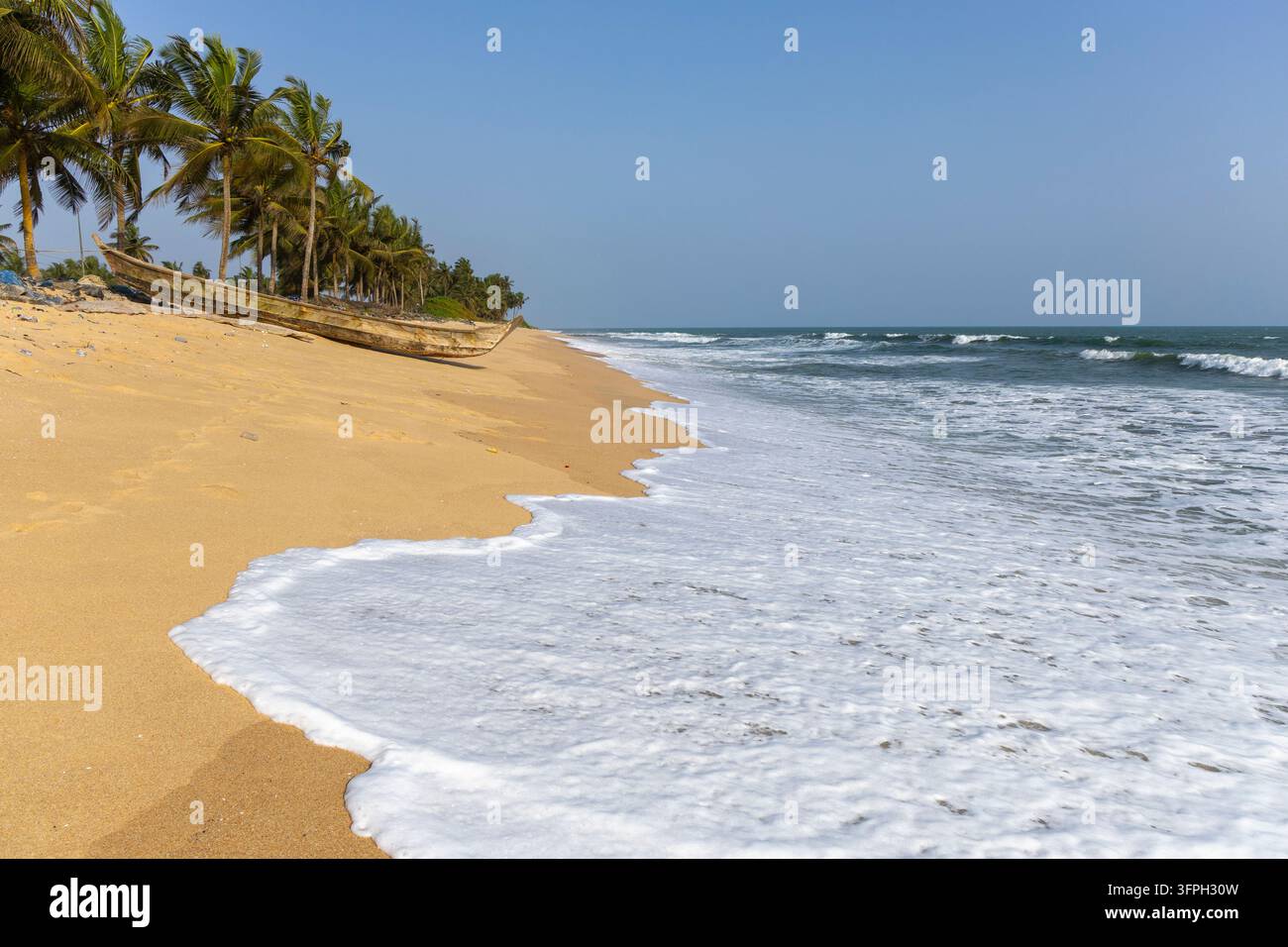 Una canoa da pesca in legno si trova su una spiaggia sabbiosa vuota fiancheggiata da palme, Regione Centrale, Elmina, Ghana Foto Stock