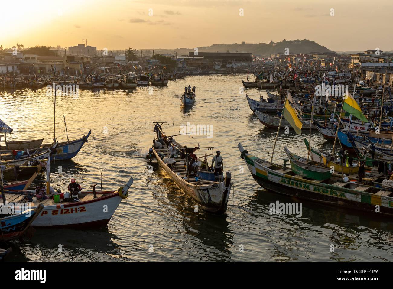 Barche di pesca in legno attraversano il canale portuale affollato all'ora dorata, Regione Centrale, Elmina, Ghana Foto Stock