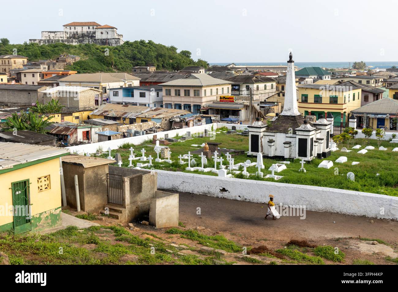 Donna con un cesto sulla testa davanti a un cimitero che si affaccia sulla città, Regione Centrale, Elmina, Ghana Foto Stock