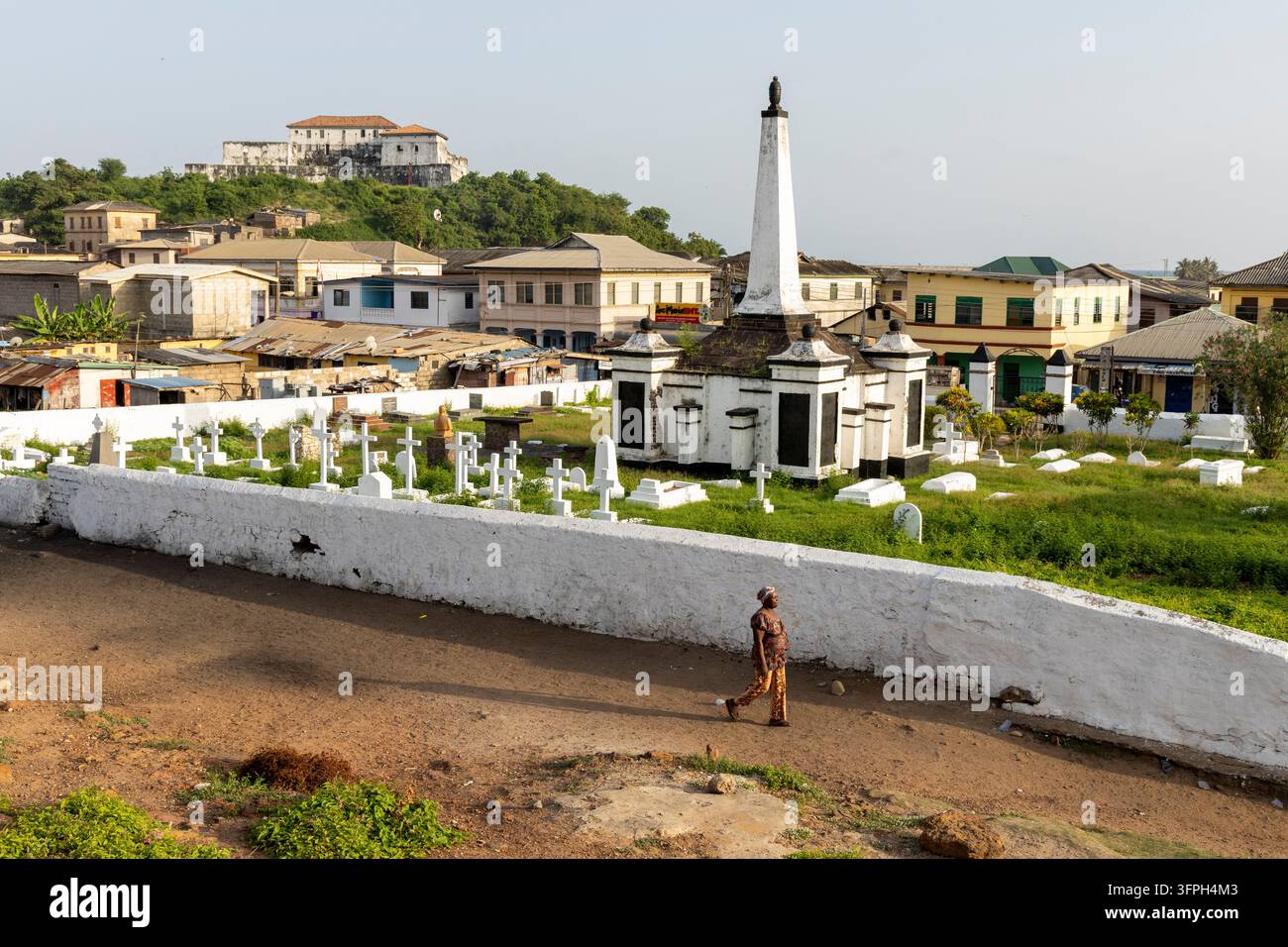 Donna che cammina lungo un sentiero oltre un obelisco del cimitero che si affaccia sulla città, Regione Centrale, Elmina, Ghana Foto Stock