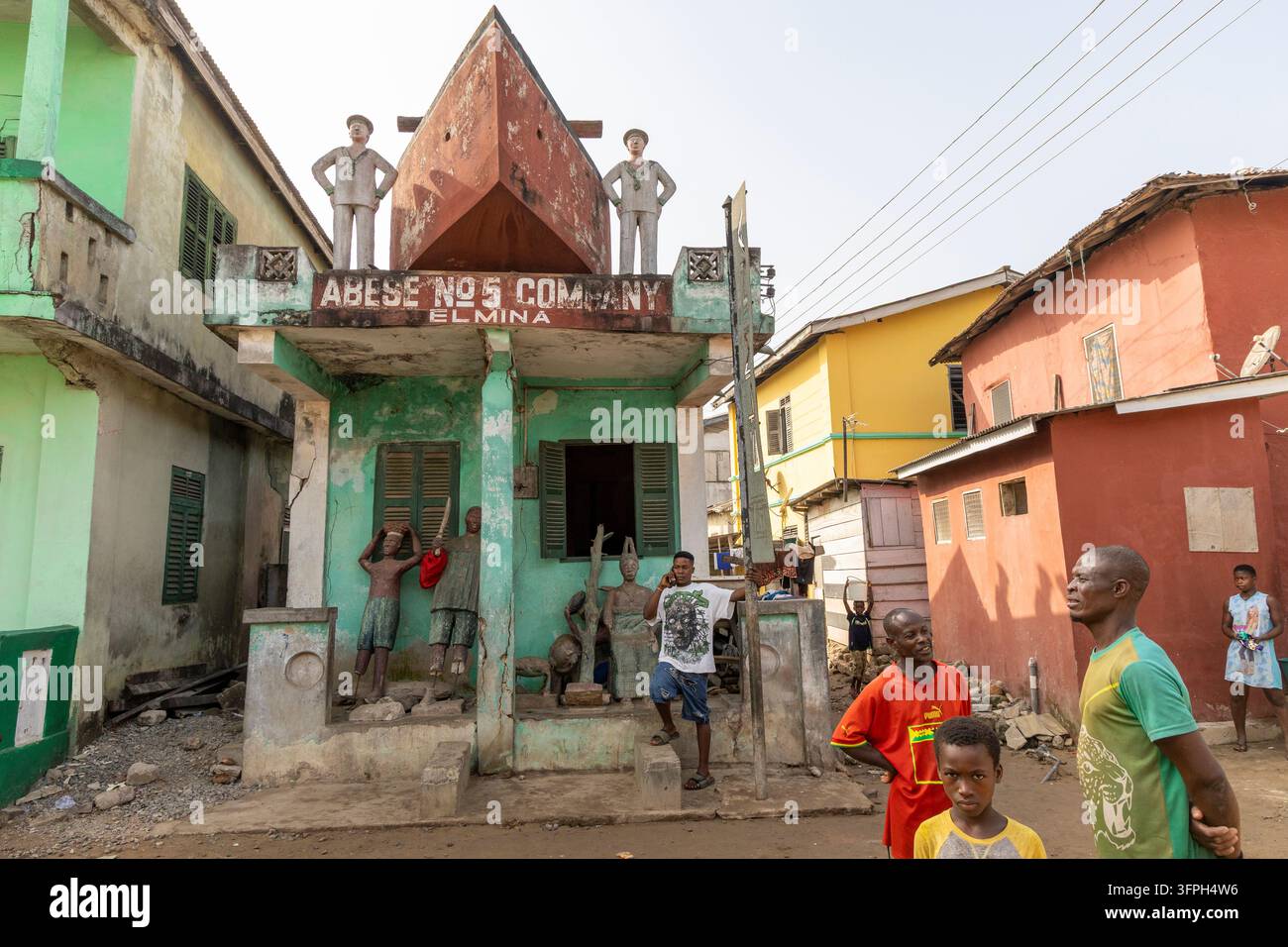 Scena di strada con residenti locali che passano davanti a una casa dipinta di una compagnia Asafo decorata con statue, Regione centrale, Elmina, Ghana Foto Stock