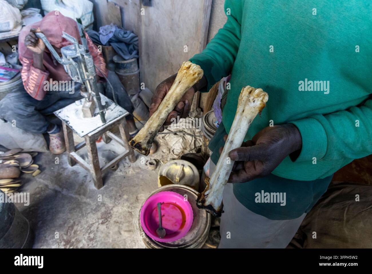 Uomo in maglione verde che mostra due ossa di mucca crude usate come materiale mentre un operaio aziona un trapano dietro di lui in un'area di souvenir in osso di Kibera Foto Stock