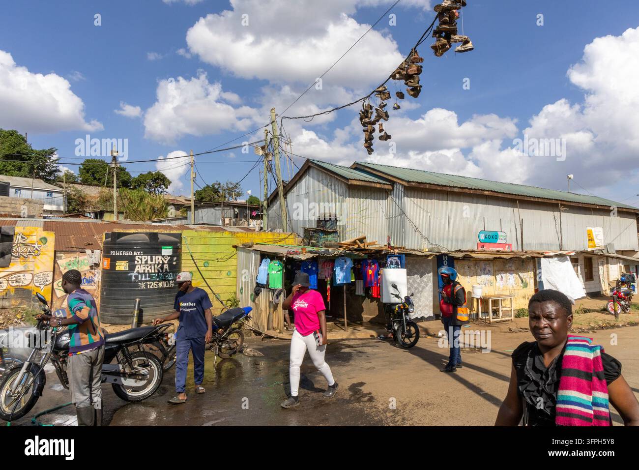 Pedoni e moto su una strada fangosa nell'area di Kibera con scarpe appese a una linea elettrica sopra, sotto un cielo blu con nuvole bianche, Nairobi Foto Stock