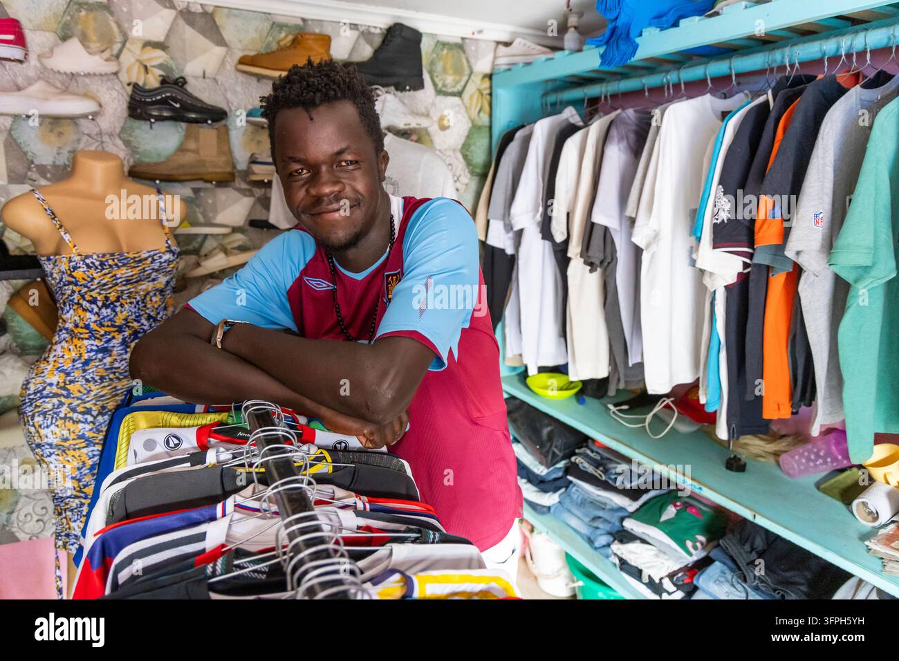 Giovane uomo sorridente in una maglia da calcio in un negozio di abbigliamento usato nell'area di Kibera, Contea di Nairobi, Nairobi, Kenya Foto Stock