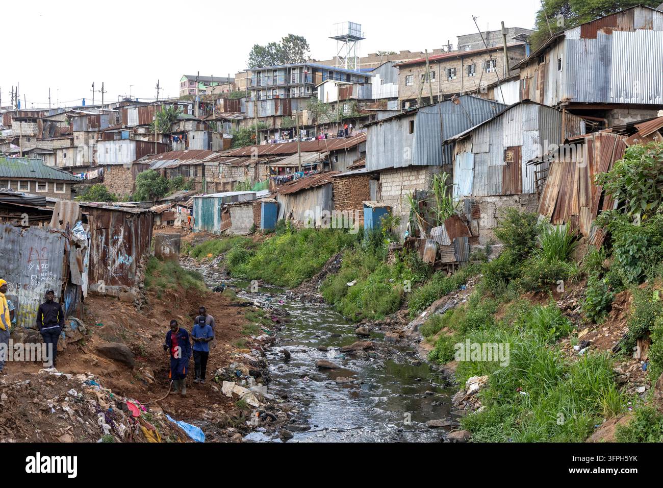 Ruscello che scorre attraverso l'area di Kibera con baracche sulla collina e uomini che camminano lungo la riva, Contea di Nairobi, Nairobi, Kenya Foto Stock