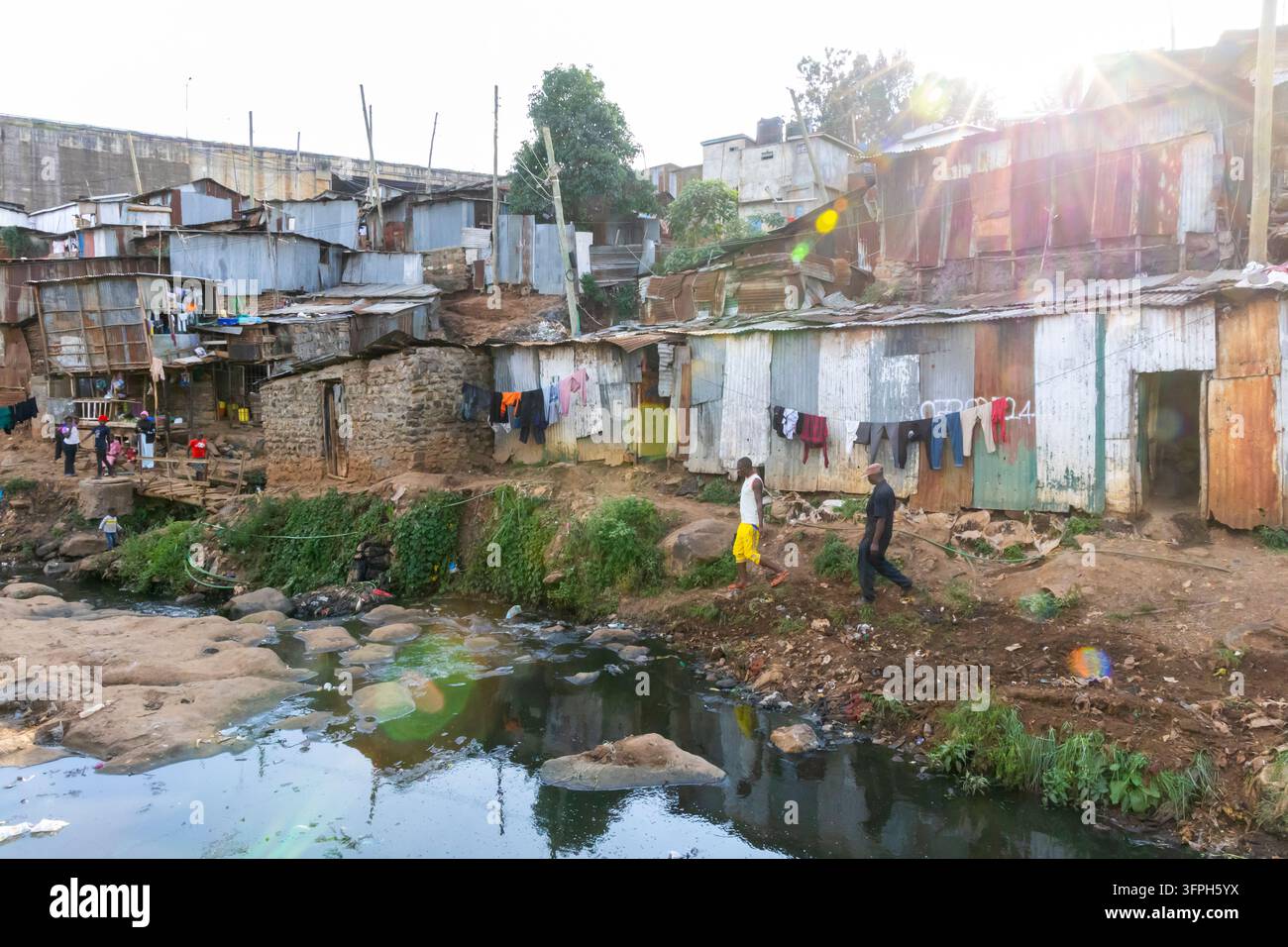 Due uomini che camminano lungo un torrente inquinato accanto a baracche di lamiera ondulata e lavanderia nell'area di Kibera, Contea di Nairobi, Nairobi, Kenya Foto Stock