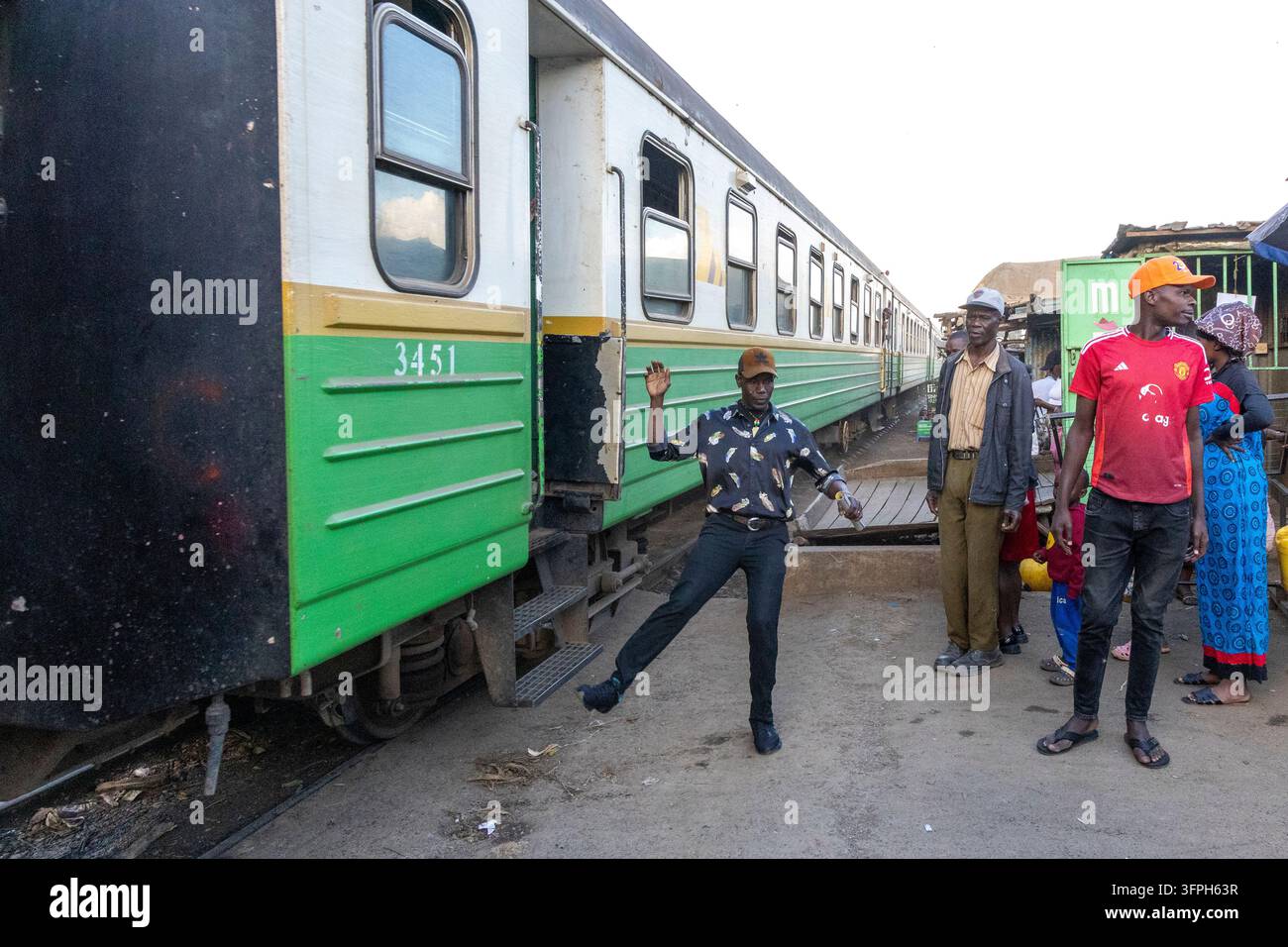Uomo con una camicia a stampa piume che balla accanto a un treno passeggeri mentre gli spettatori osservano nell'area di Kibera, Contea di Nairobi, Nairobi, Kenya Foto Stock