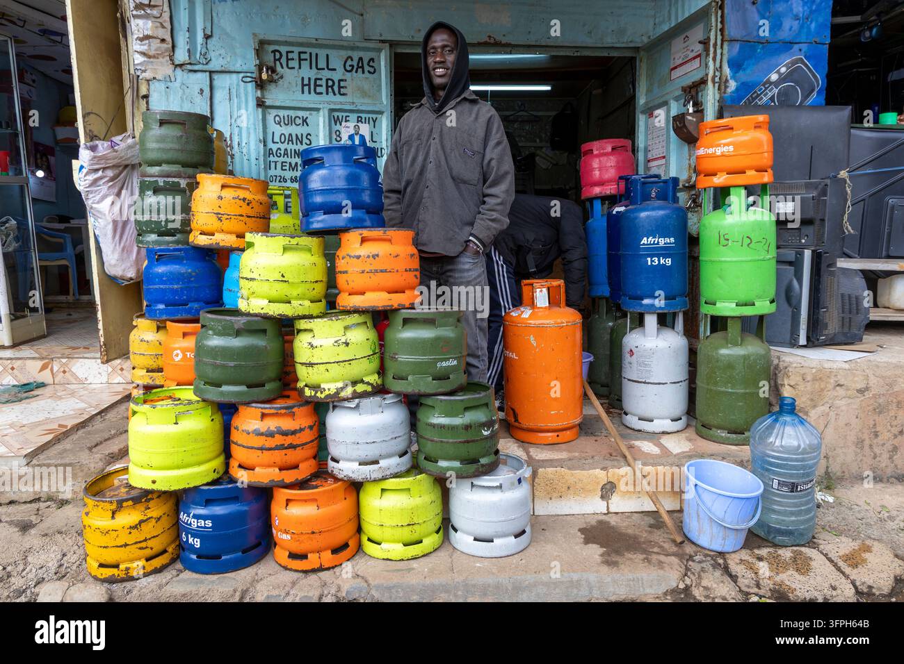 Uomo sorridente con giacca con cappuccio in piedi dietro bombole di gas colorate impilate in un negozio di rifornimento di gas nell'area di Kibera, Contea di Nairobi, Nairobi, Kenya Foto Stock