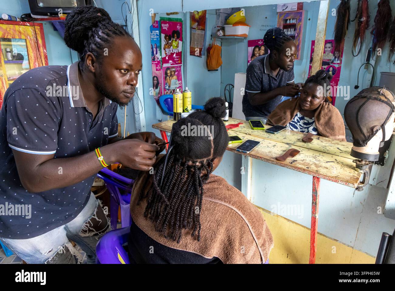 Parrucchiere che acconcia i capelli di una donna con un pettine in un piccolo salone con uno specchio nell'area di Kibera, Contea di Nairobi, Nairobi, Kenya Foto Stock