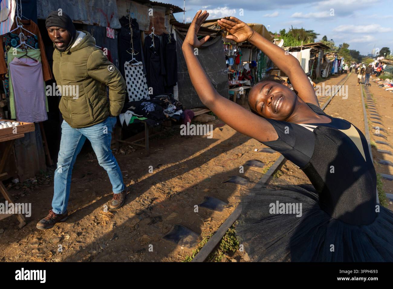 Ballerina sorridente in tutù nero che si inarca con le braccia alzate sui binari ferroviari della scuola di balletto di Kibera mentre un uomo con una giacca verde passa oltre Foto Stock