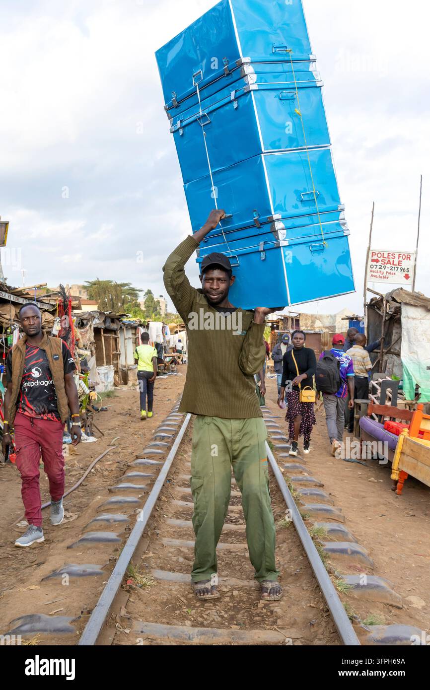 Uomo che porta una pila di bauli metallici blu sulla testa mentre cammina lungo i binari ferroviari attraverso l'area di Kibera con bancarelle del mercato e pedoni, N Foto Stock