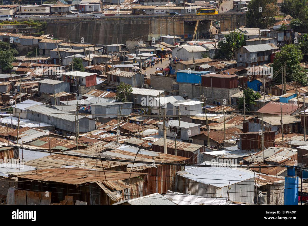 Vista sulle dense tettoie di lamiera ondulata dell'area di Kibera con una strada trafficata e un bus giallo che attraversa un ponte sullo sfondo, Contea di Nairobi Foto Stock
