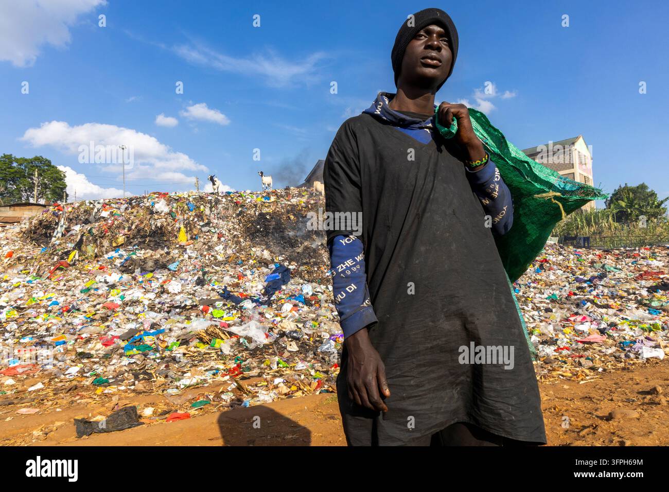 Giovane uomo con un berretto nero che porta un sacco verde sulla spalla davanti a un enorme cumulo di rifiuti con capre in cima nella zona di Kibera, Contea di Nairobi Foto Stock