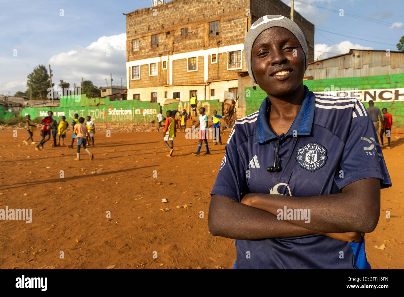 Allenatrice sorridente per una squadra di calcio a Kibera con le braccia incrociate mentre dei ragazzi giocano su un campo di terra dietro di lei, Contea di Nairobi, Nairobi, Kenya Foto Stock