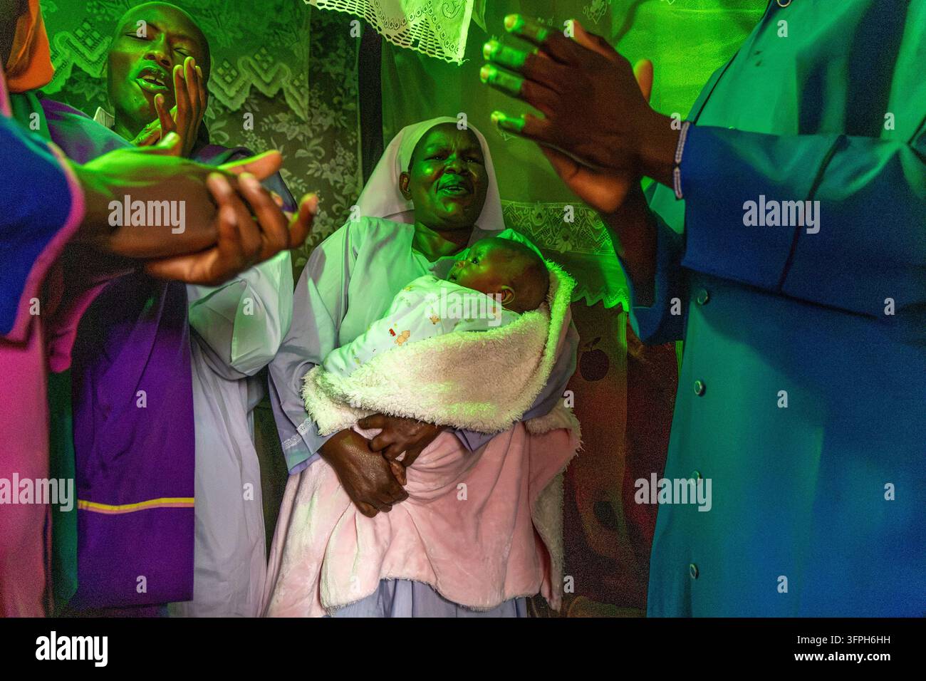 Donna in velo bianco che tiene un neonato mentre i fedeli applaudono e cantano durante un battesimo a Kibera sotto luce verde, Contea di Nairobi, Nairobi, Kenya Foto Stock