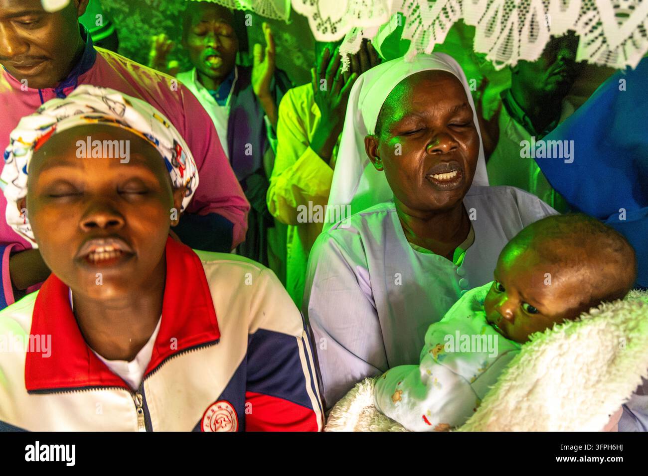 Donne che pregano a occhi chiusi mentre una con un velo bianco tiene un bambino durante un battesimo a Kibera, Contea di Nairobi, Nairobi, Kenya Foto Stock