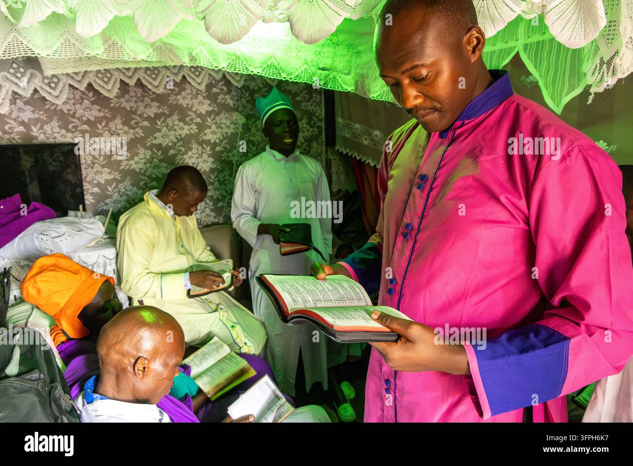 Uomo in una veste rosa e blu che legge una Bibbia mentre altri uomini leggono nelle vicinanze durante un battesimo a Kibera, Contea di Nairobi, Nairobi, Kenya Foto Stock
