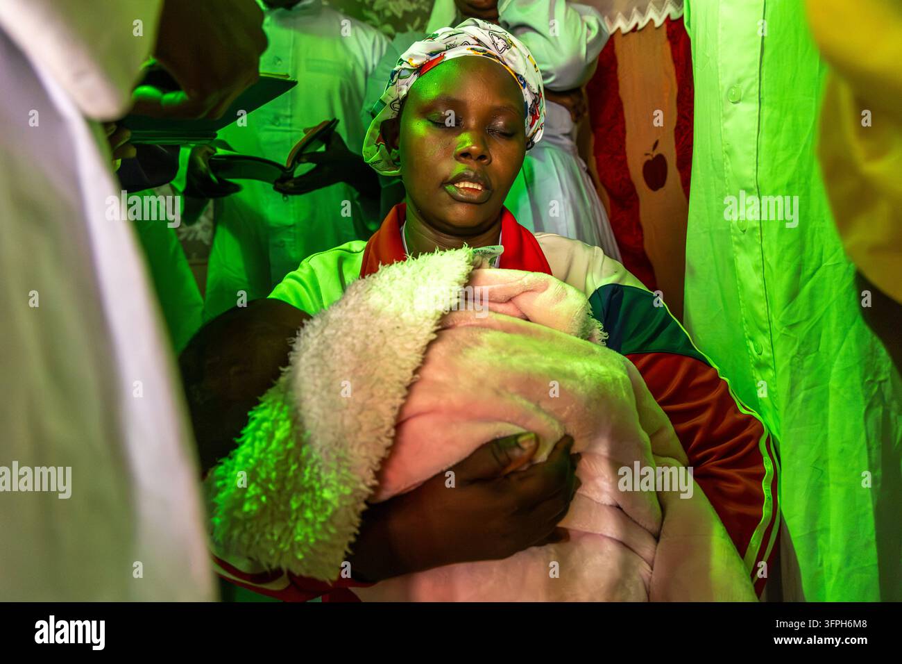 Donna con foulard stampato e occhi chiusi che tiene un bambino avvolto in una coperta rosa durante un battesimo a Kibera, Contea di Nairobi, Nairobi, Kenya Foto Stock