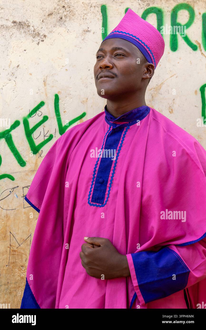 Uomo in una veste rosa e blu e cappello abbinato in piedi davanti a un muro di graffiti nell'area di Kibera, Contea di Nairobi, Nairobi, Kenya Foto Stock