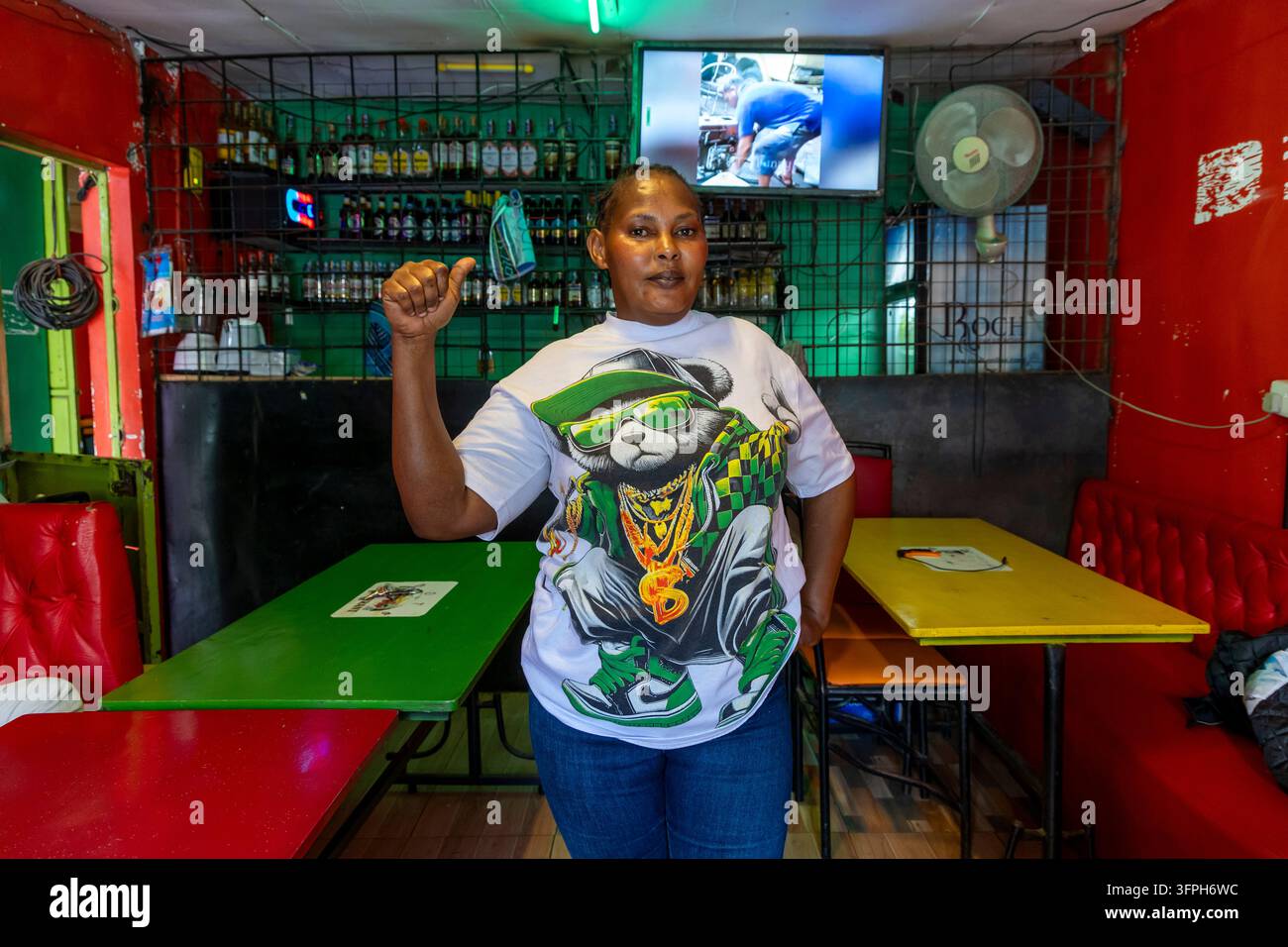 Donna in un bar nella zona di Kibera che fa il pollice alzato in una maglietta con un grafico di un orso davanti a tavoli colorati e bottiglie, Contea di Nairobi, Nairobi, Ke Foto Stock