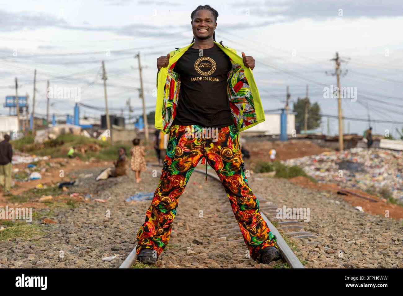 David Avido, stilista di moda, sorride e apre il suo blazer verde lime per mostrare una maglietta Made in Kibra mentre è in piedi su una linea ferroviaria con un rifiuto Foto Stock