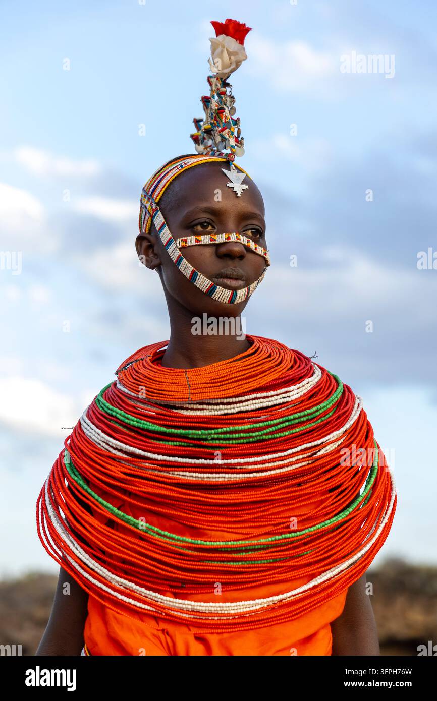 Ritratto di una donna della tribù Samburu con una fascia per il viso con perline, un pendente sulla fronte in argento, un copricapo floreale e un enorme collare di perle rosse su uno sfondo di cielo blu Foto Stock