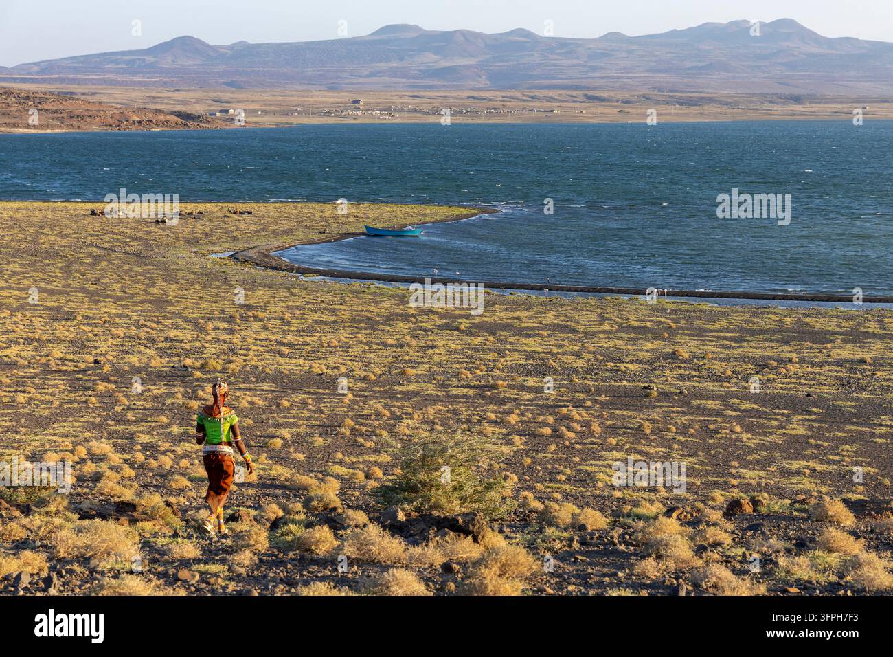 Donna del tribù Rendille in una maglia verde e copricapo di perline che cammina su una riva erbosa verso il lago Turkana con una barca blu e montagne in lontananza, Marte Foto Stock