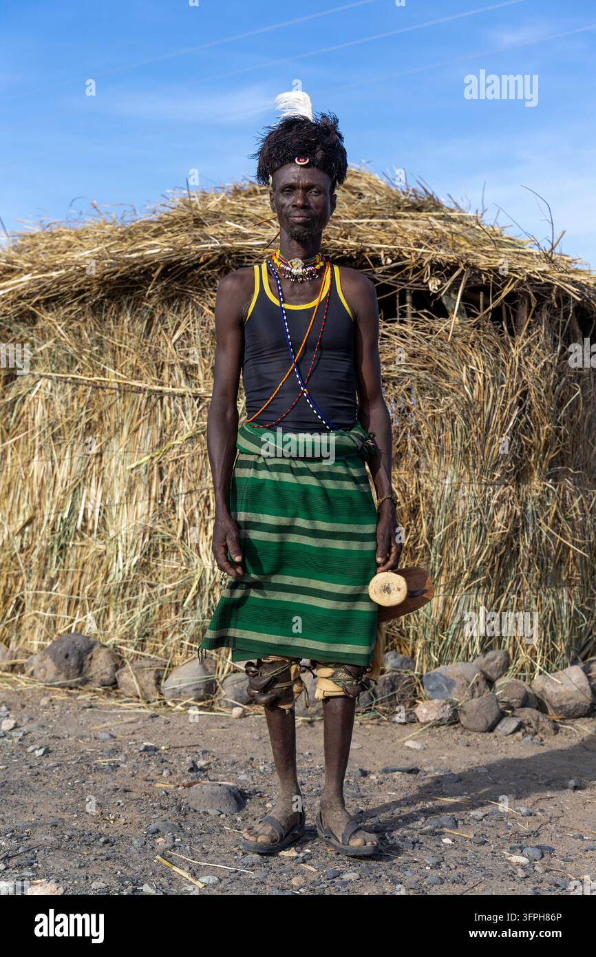 Uomo in piedi della tribù Turkana con copricapo di piume e gonna verde che tiene un supporto per la testa in legno davanti a una capanna, Contea di Marsabit, Loiyangalani, Kenya Foto Stock