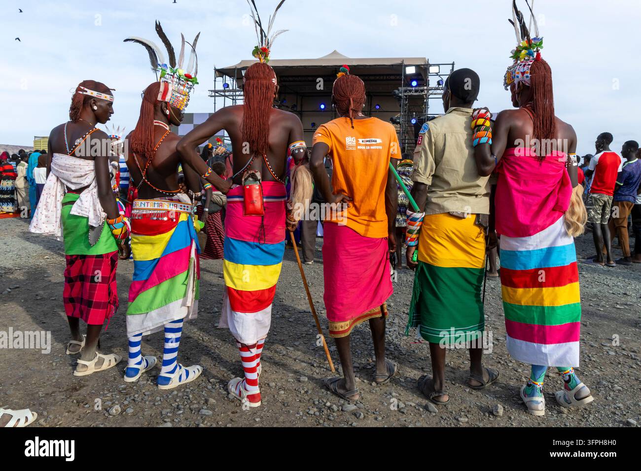 Vista posteriore di uomini della tribù Rendille con capelli intrecciati con ocra rossa, copricapo piumato e fasce colorate che osservano un palco al Festival Culturale del Lago Turkana Foto Stock
