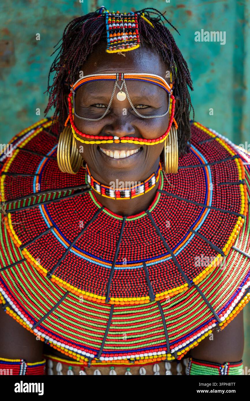Ritratto ravvicinato di una donna sorridente della tribù Pokot con un velo facciale di perline e orecchini a catena in ottone e un ampio colletto di perline rosse e verdi davanti a un turchese Foto Stock