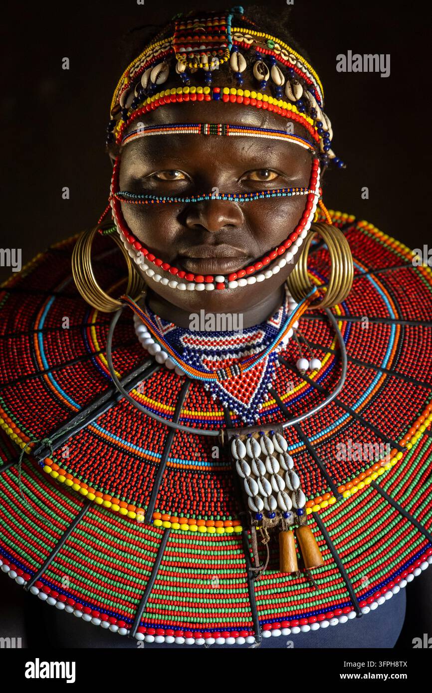 Ritratto ravvicinato di una donna della tribù Pokot che indossa un enorme collare di perle e un copricapo in conchiglie di cowrie con cinghie perlate sul viso e orecchini di ottone in un ambiente scuro Foto Stock
