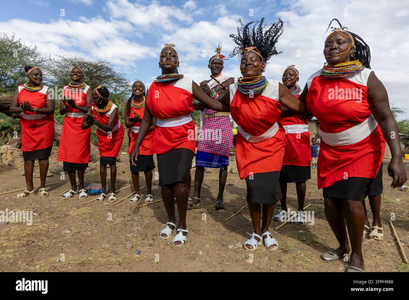 Donne della tribù Njemps che danzano in abiti rossi con collane di perle e fasce per il viso mentre un uomo in un tessuto a quadri si trova dietro di loro, Lago Baringo, Kenya Foto Stock