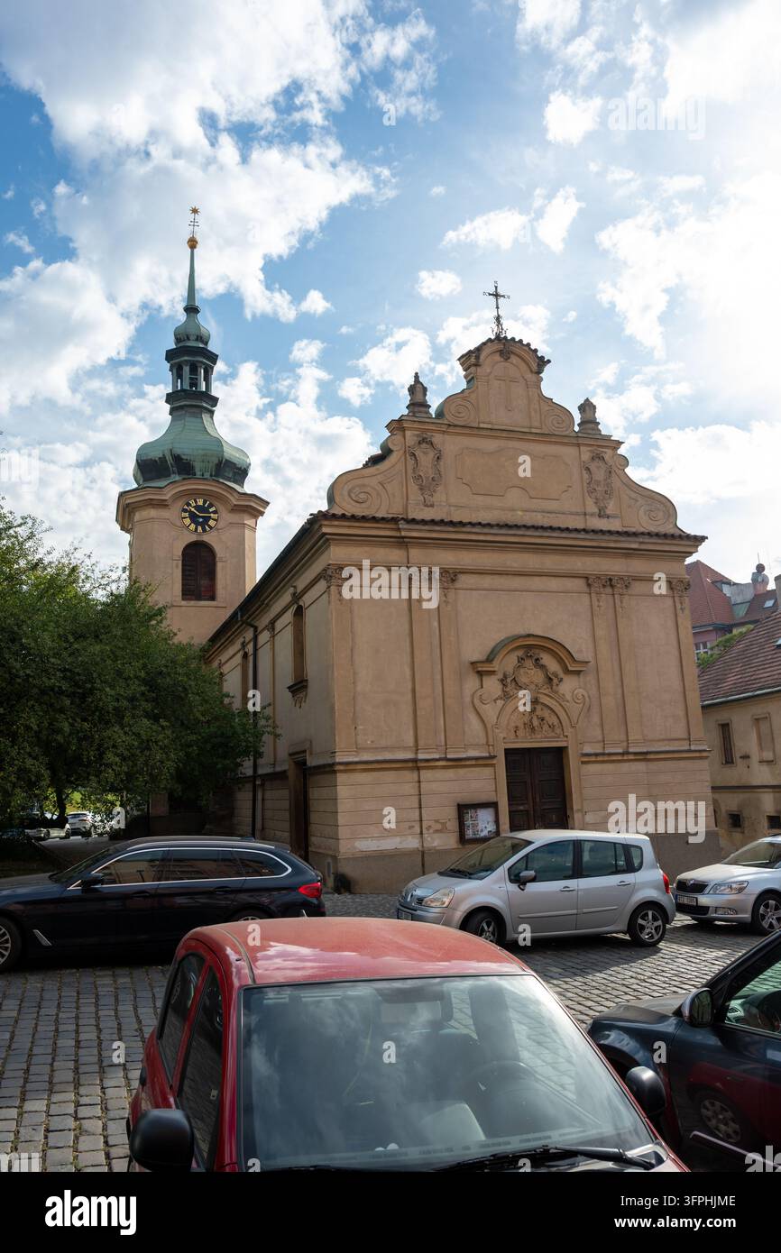 Una chiesa barocca storica con una facciata ornata, torre dell'orologio e punta in rame verde, circondata da auto parcheggiate su una strada acciottolata. Foto Stock