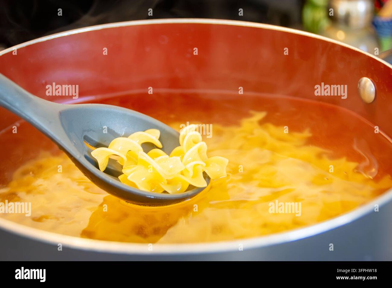 Preparazione del pasto Pollo Alfredo facile: larghe tagliatelle alle uova che bollono in una pentola rossa con un cucchiaio a fessura che raccoglie la pasta cotta. Foto Stock