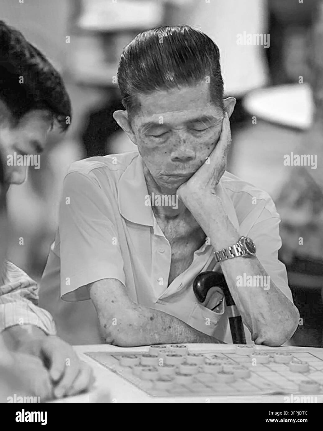 Fotografia documentaria in bianco e nero di un anziano uomo concentrato su un gioco da tavolo cinese tradizionale a Chinatown, Singapore, appoggiando il viso a Foto Stock