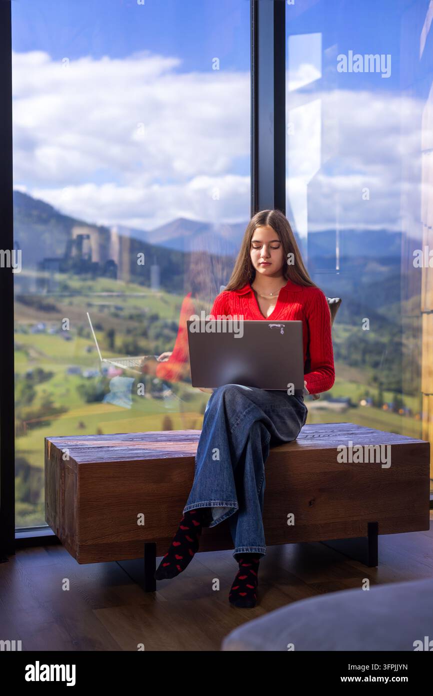 Giovane donna che lavora su un laptop accanto a una finestra panoramica, lavoro da remoto in un moderno rifugio di montagna con una valle e montagne scenografiche sullo sfondo. Foto Stock