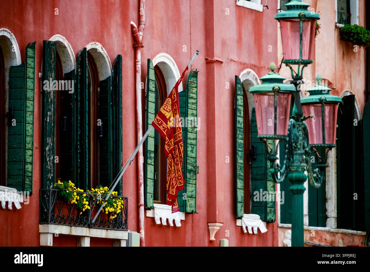 Edificio colorato sul lato del canale veneziano con pareti rosse sbiadite, persiane verdi, finestre ad arco, fioriere, una bandiera decorativa e lampioni vintage. Foto Stock