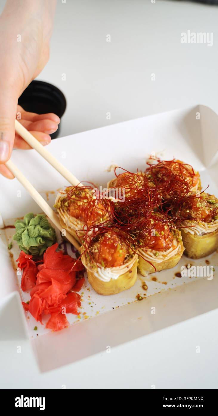 Mano che prende con le bacchette un rotolo di sushi fusion da una scatola da asporto, godendosi la moderna cucina giapponese consegnata a domicilio Foto Stock