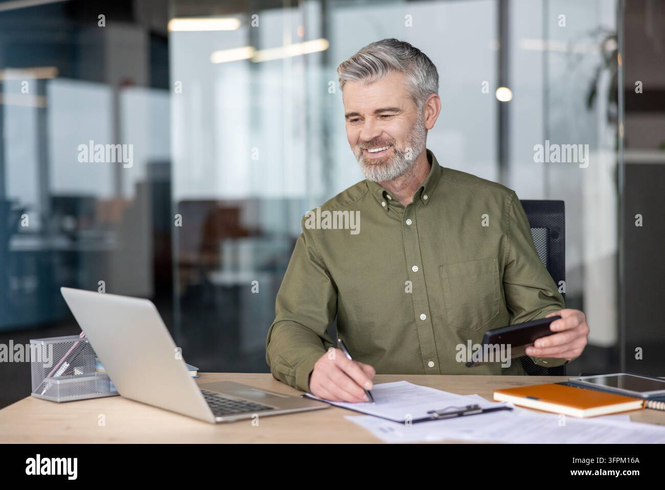 Uomo d'affari maturo che calcola le finanze utilizzando una calcolatrice, rivede documenti e lavora su un laptop alla sua scrivania in ufficio, simboleggiando la contabilità professionale e la gestione finanziaria Foto Stock