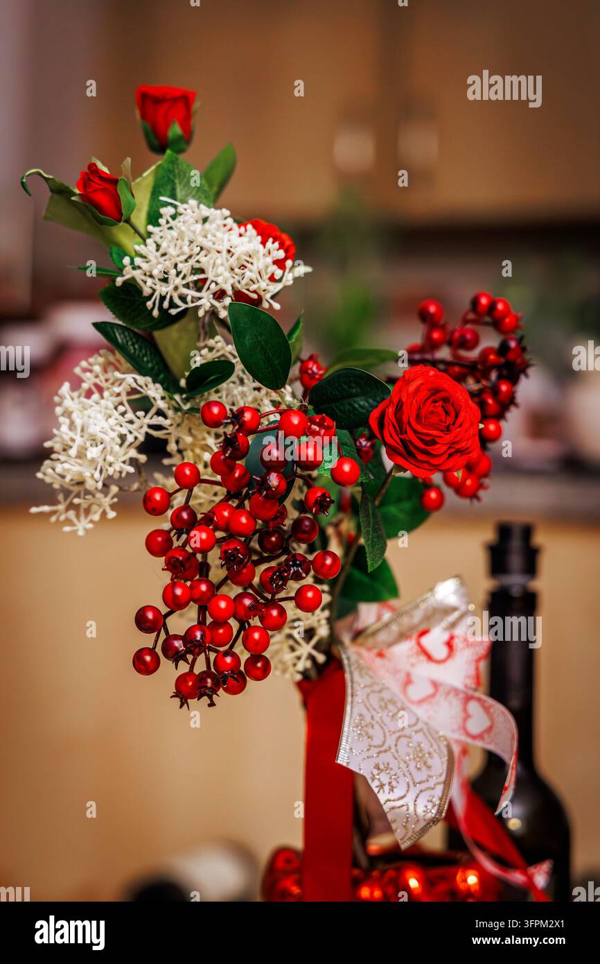 Un primo piano mostra un bellissimo mazzo di rose rosse, fiori bianchi delicati e bacche rosse brillanti disposti in un vaso decorativo legato con un f Foto Stock