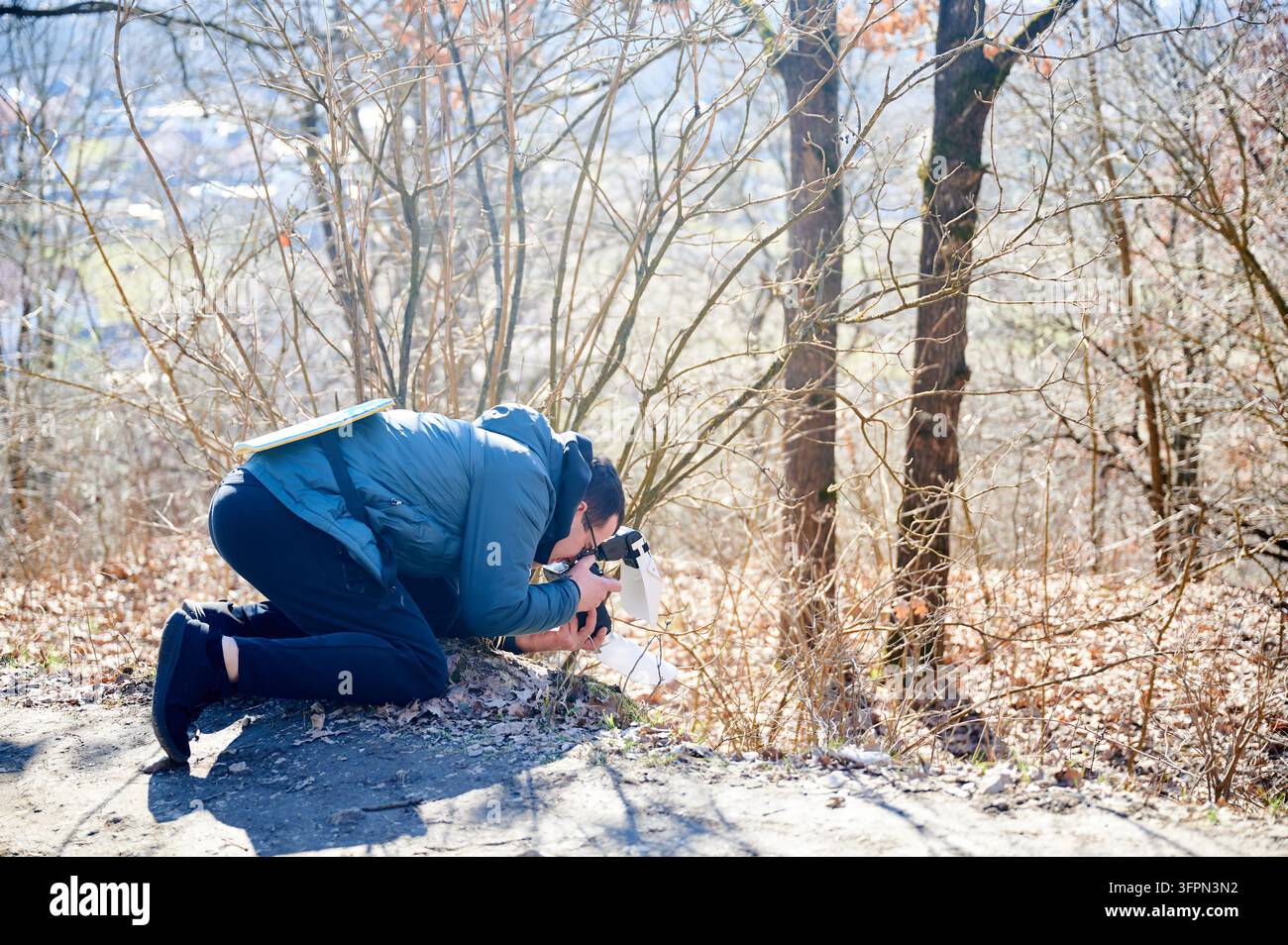 Fotografo outdoor si inginocchia su un sentiero boschivo per catturare una scena naturale in primo piano con la fotocamera nella luce del primo primavera. Utile per la promozione turistica, marchi outdoor, marketing del tempo libero e concetti di stile di vita Foto Stock