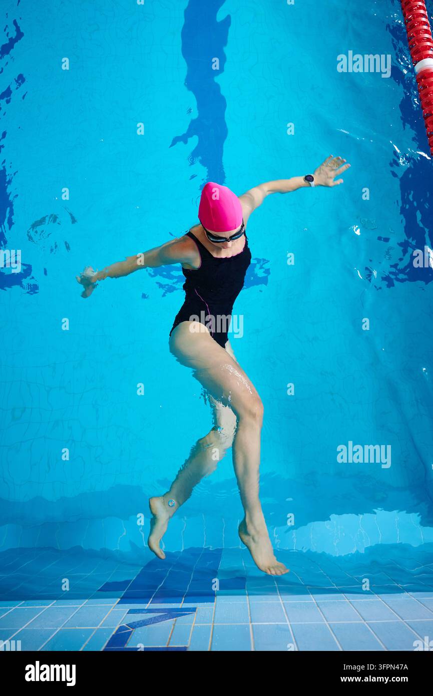 Giovane nuotatrice sott'acqua nella piscina, muovendo braccia e gambe, indossa costume da bagno, cuffia e occhialini, pratica allenamento acquatico e esercizi di fitness Foto Stock