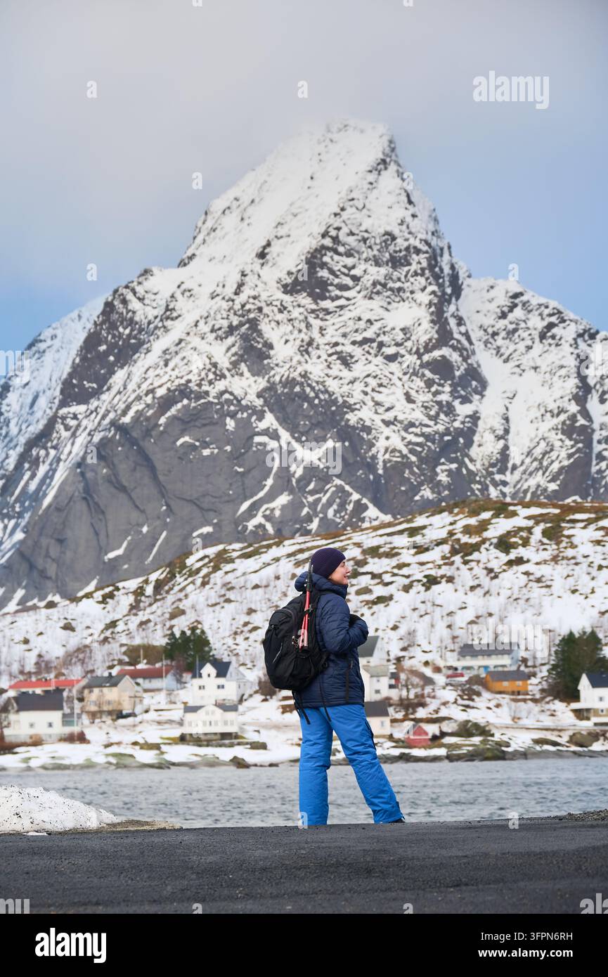Donna viaggiatrice cammina con zaino e treppiede accanto al Reinefjorden, incorniciata dall'Olstinden ricoperto di neve e dalle case invernali di Reine, Isole Lofoten, Norvegia Foto Stock