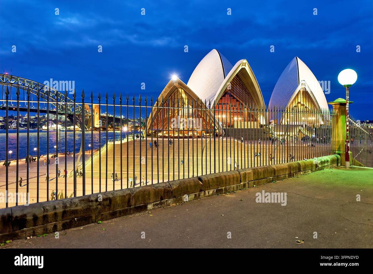 I visitatori percorrono i gradini del cortile antistante sotto le vele illuminate di Bennelong Point all'ora blu, con il Harbour Bridge sullo sfondo, NSW, Australia Foto Stock