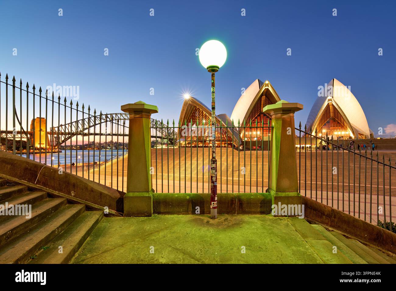 Gradini in arenaria, ringhiere in ferro e un lampione incorniciano il luogo di concerto illuminato su Bennelong Point, New South Wales, Australia, al crepuscolo. Foto Stock