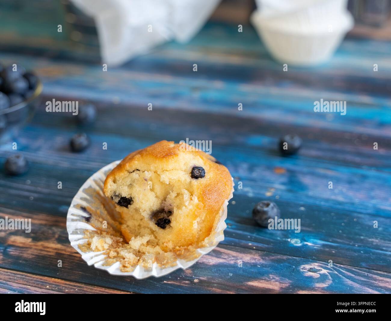 Vista ravvicinata di un muffin ai mirtilli appena sfornato con mirtilli freschi sparsi su una superficie in legno blu invecchiato e un accessorio da forno sfocato in morbido Foto Stock