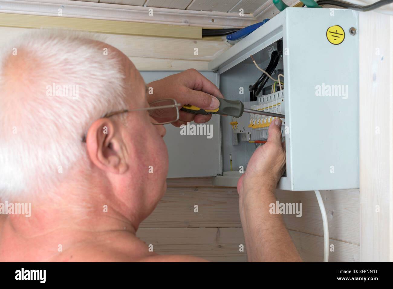 Elettricista anziano stringe con attenzione una vite nel quadro dei fusibili, concentrandosi sulla precisione e la sicurezza durante la manutenzione elettrica a casa. Foto Stock