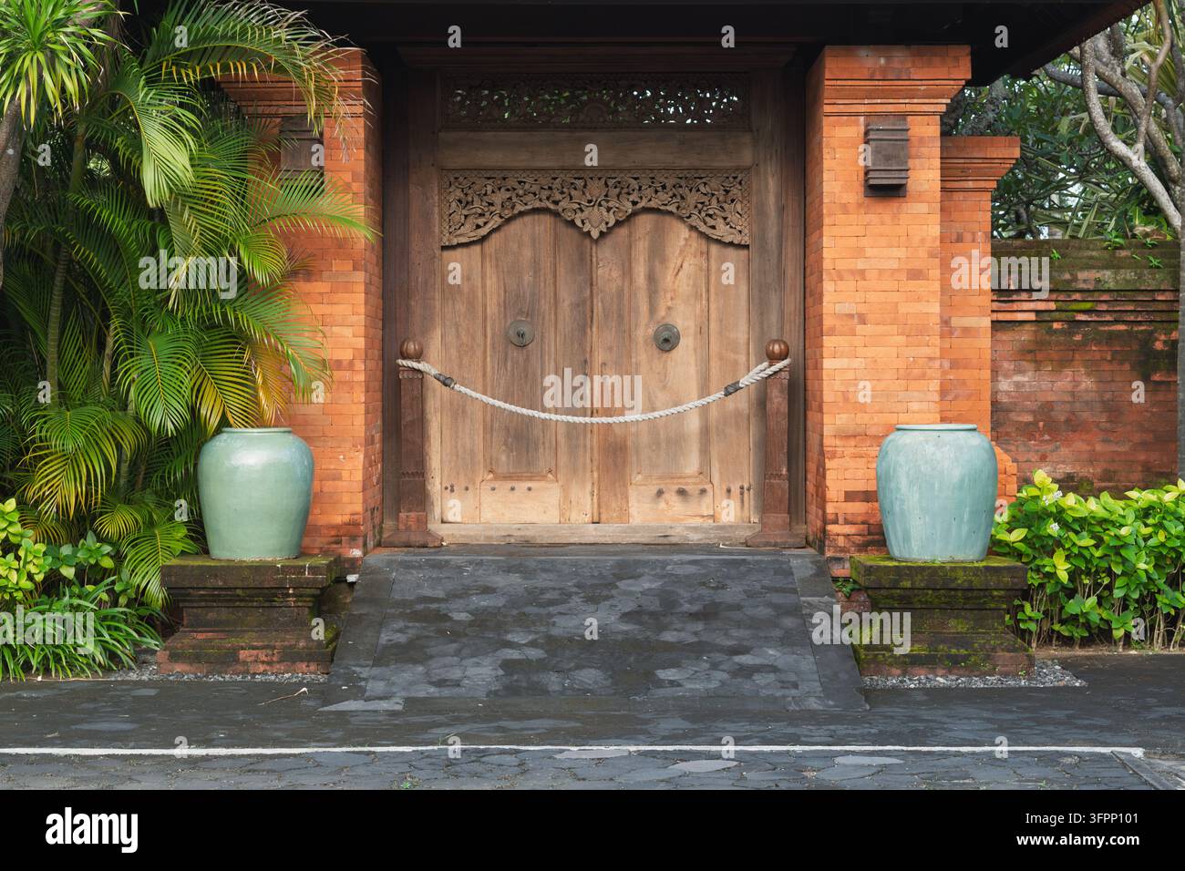 Cancello di legno intagliato chiuso con barriera in corda si trova tra muri di mattoni rossi e piante tropicali all'ingresso tranquillo di una proprietà a Bali. Foto Stock