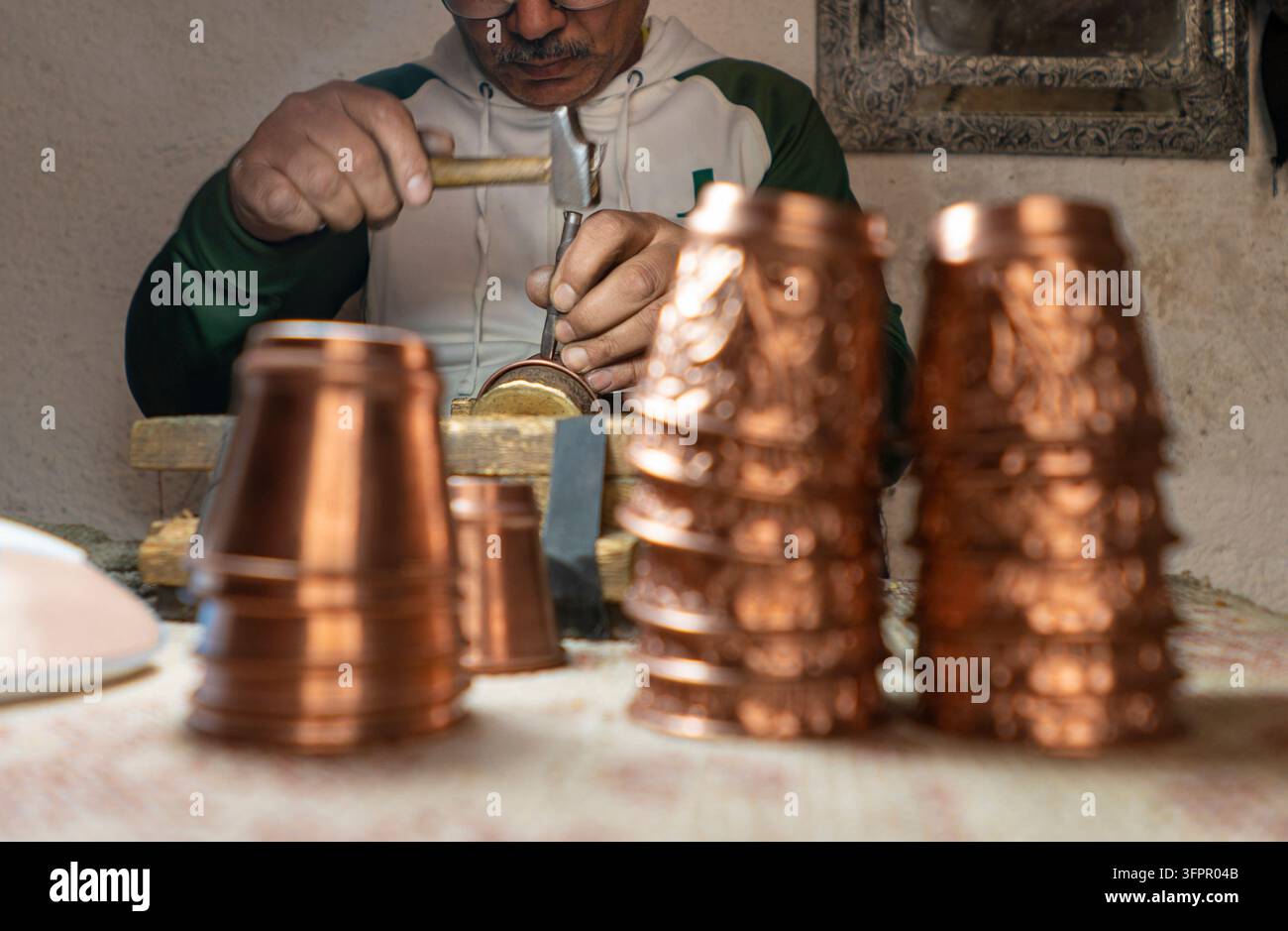 Artigiano esperto che incide disegni decorativi su una tazza di rame usando un martello e uno scalpello, con pile di tazze di rame finite in primo piano Foto Stock